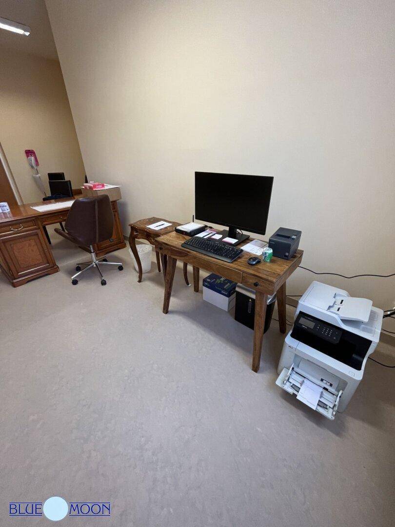 Büroarbeitsplatz mit Computer, Drucker und einem weiteren Schreibtisch im Hintergrund.