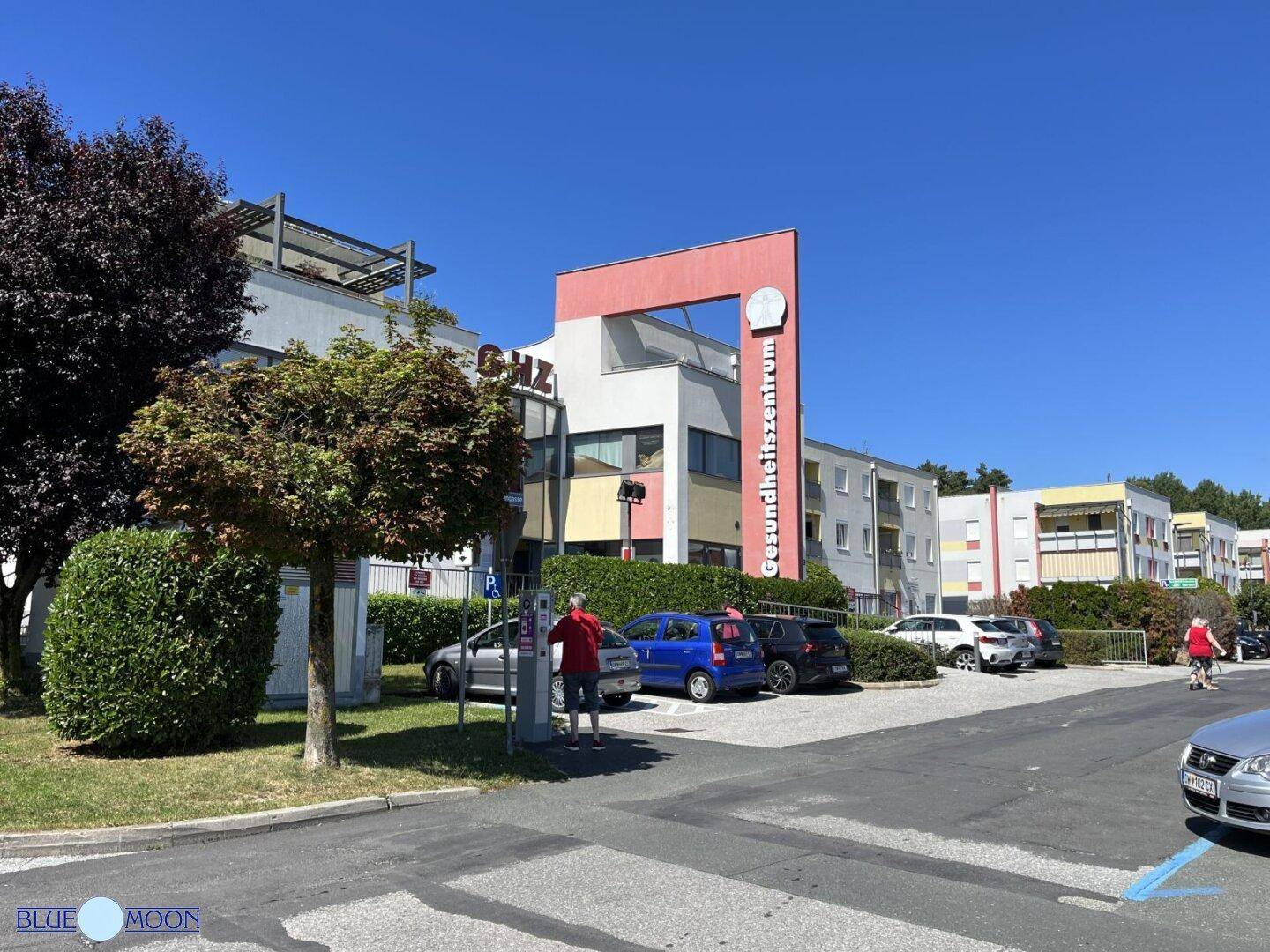 Modernes Gesundheitszentrum mit Parkplätzen und gepflegter Außenanlage an einem sonnigen Tag.