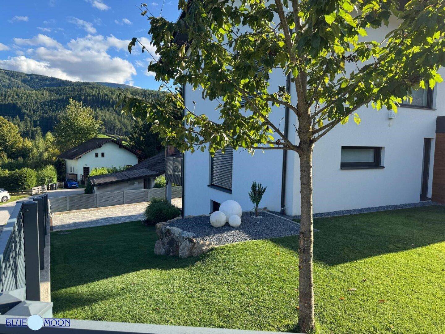 Sonnige Rasenfläche im Vorgarten mit dekorativen Elementen und Blick auf die Berge.