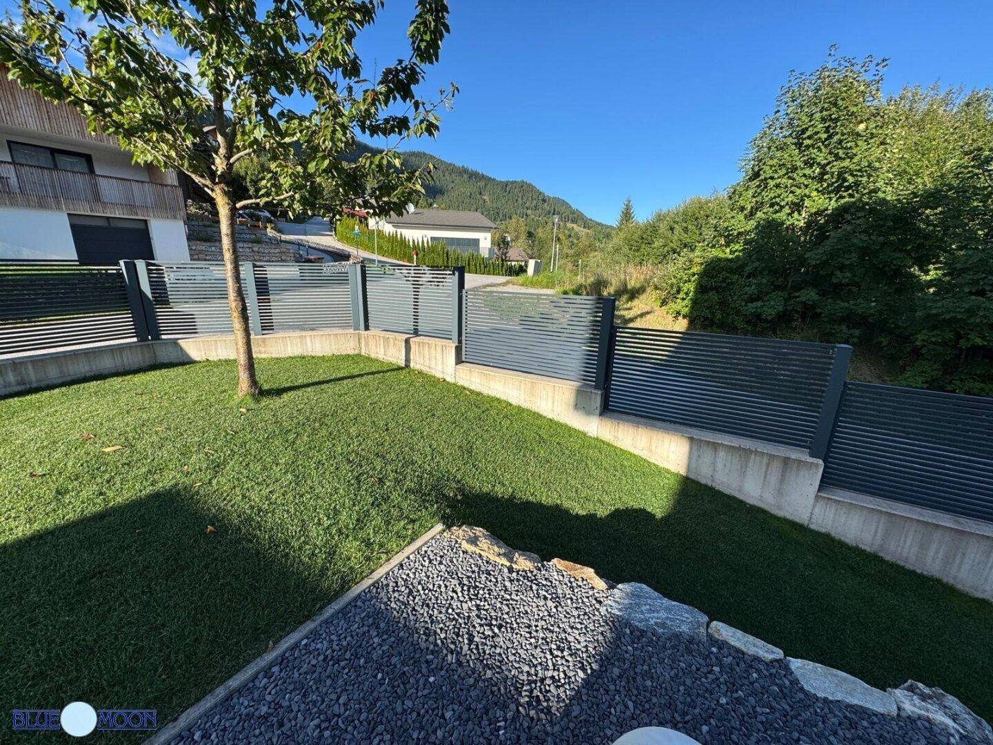 Gepflegter Gartenbereich mit Steinen, Rasen und Zaun, umgeben von grüner Landschaft.