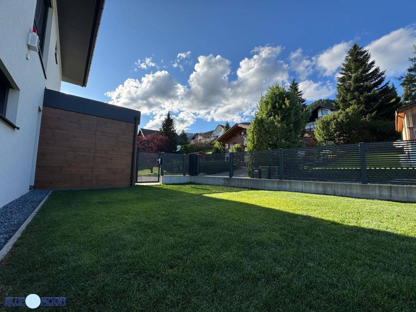 Weitläufiger Gartenbereich mit Rasen und Zaun, umgeben von Bäumen und Häusern.