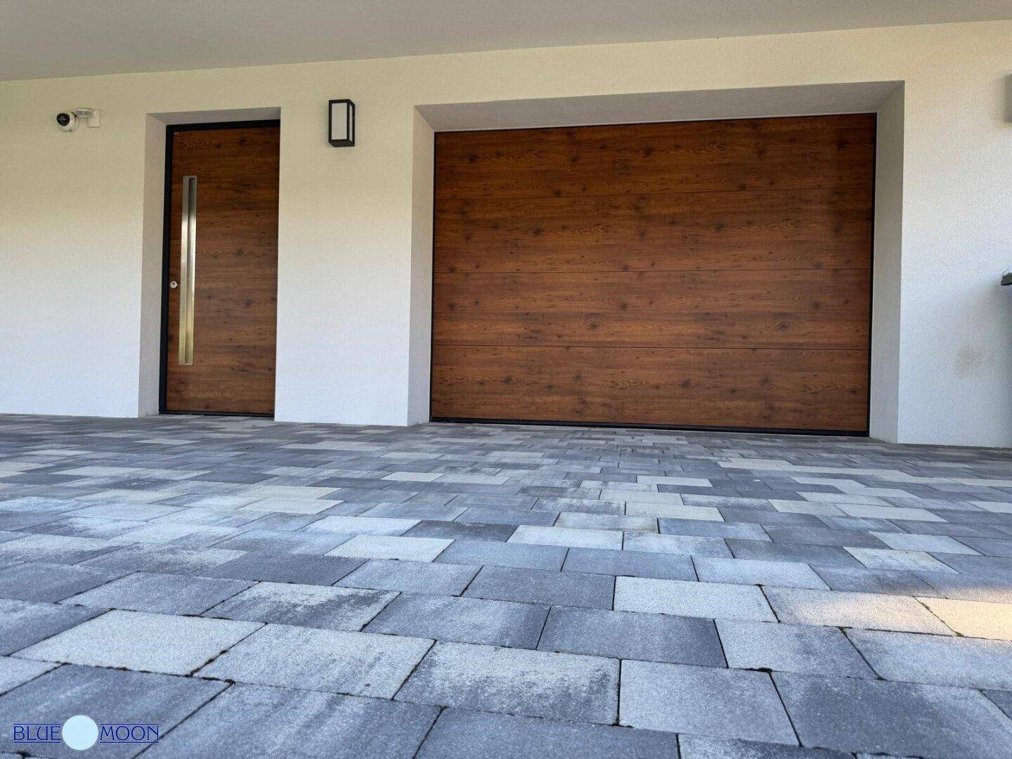 Moderne Garagentür und Eingangstür aus Holz mit gepflastertem Vorplatz.