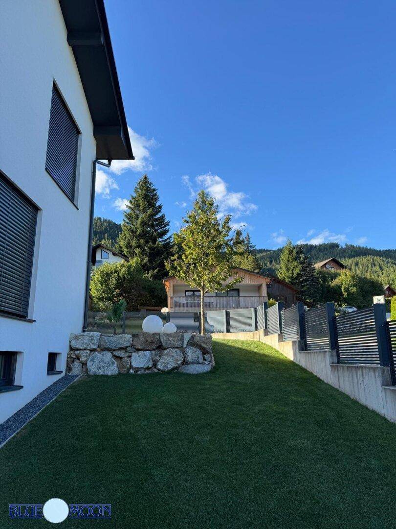 Abfallende Rasenfläche mit dekorativen Steinen und einem jungen Baum vor dem Haus.