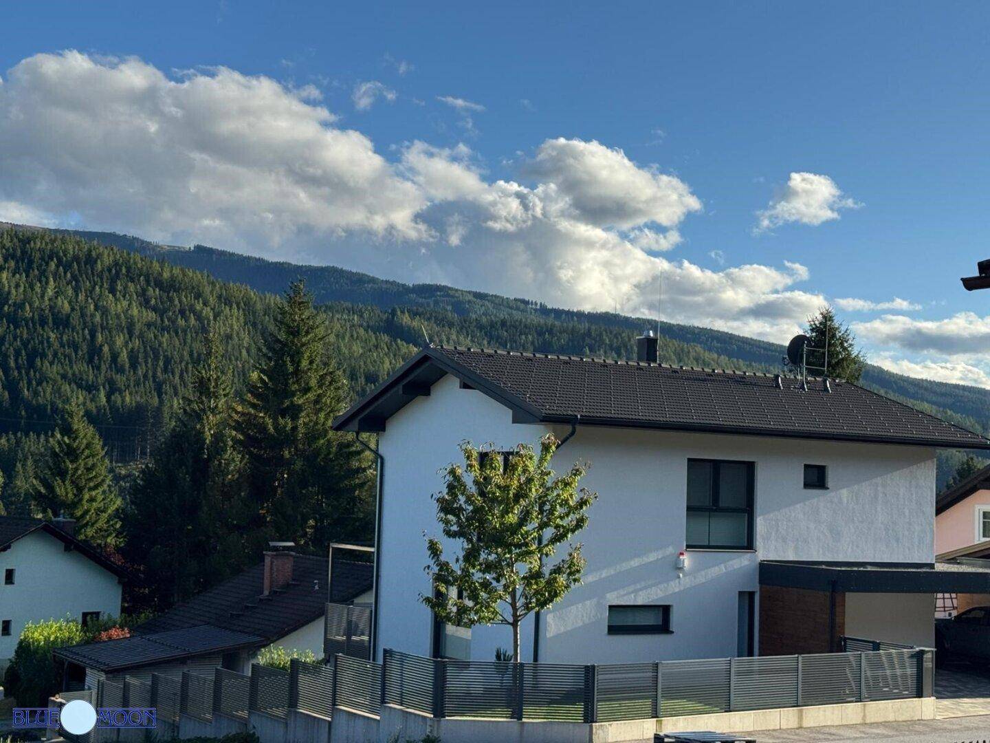 Modernes Einfamilienhaus mit weißer Fassade und dunklem Dach vor einer Bergkulisse.