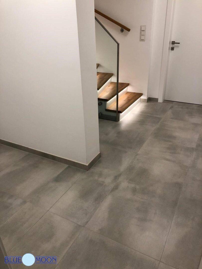Moderner Eingangsbereich mit gefliestem Boden und offener Holztreppe mit Glasgeländer.