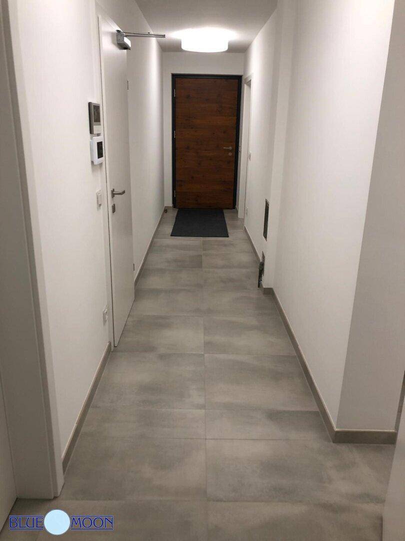 Langer, heller Flur mit grauen Fliesen und Holzeingangstür am Ende.