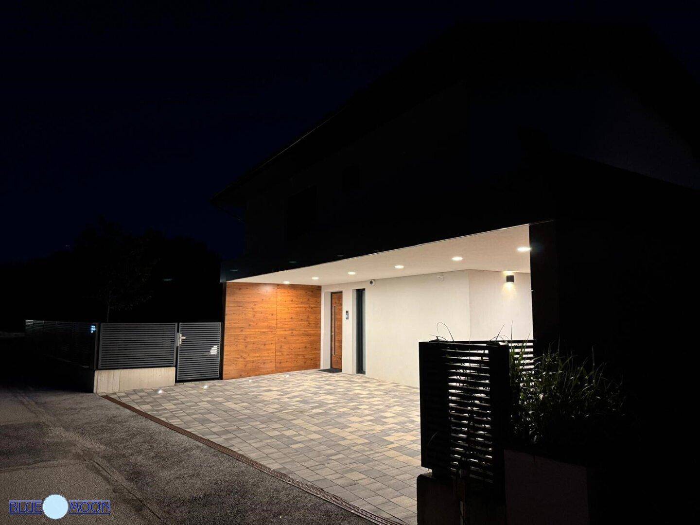 Moderne Hausfassade bei Nacht mit beleuchtetem Eingangsbereich und überdachtem Carport.