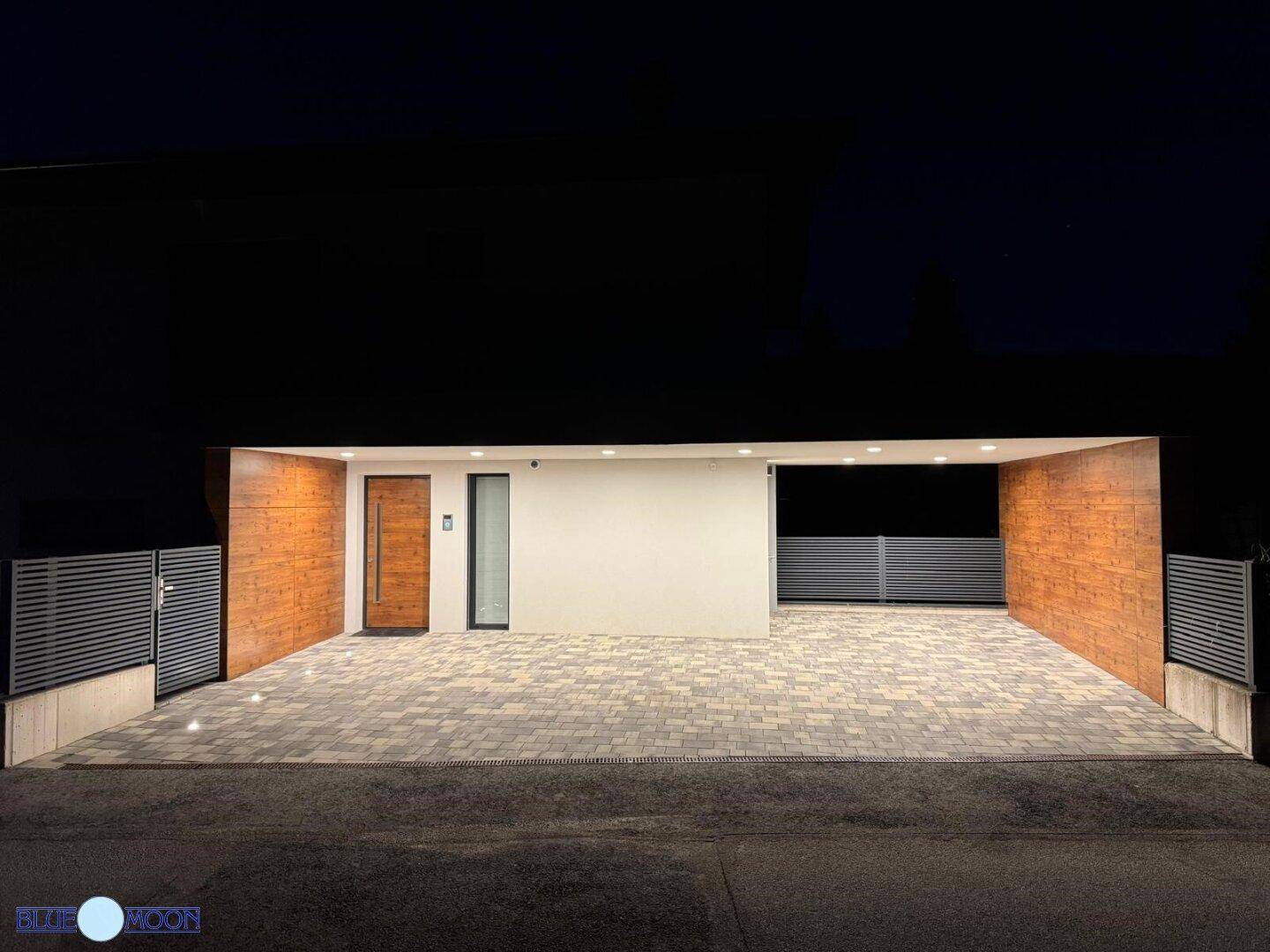 Beleuchteter Eingangsbereich und Carport eines modernen Hauses in der Nacht mit Holzakzenten.