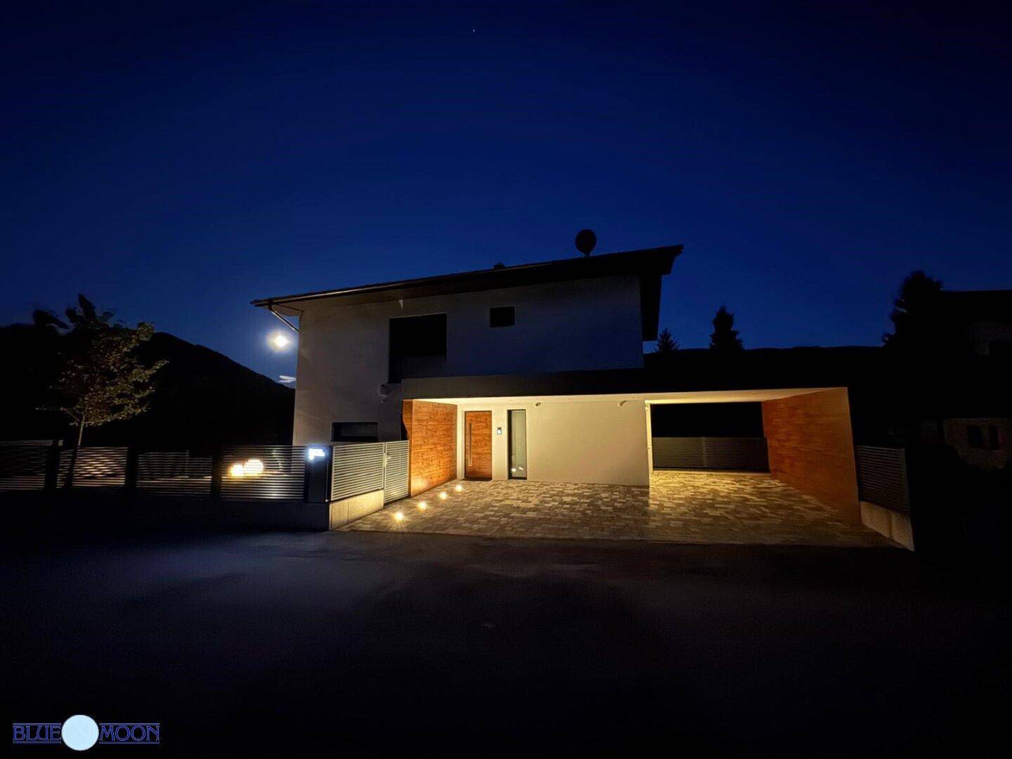 Modernes Haus bei Nacht mit beleuchteter Einfahrt und Bergblick unter klarem Nachthimmel.