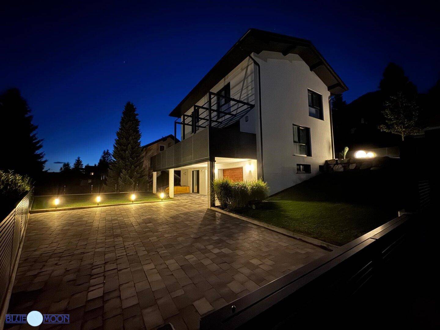 Modernes Haus bei Nacht mit beleuchteter Einfahrt, Garten und Bergblick in ruhiger Lage.