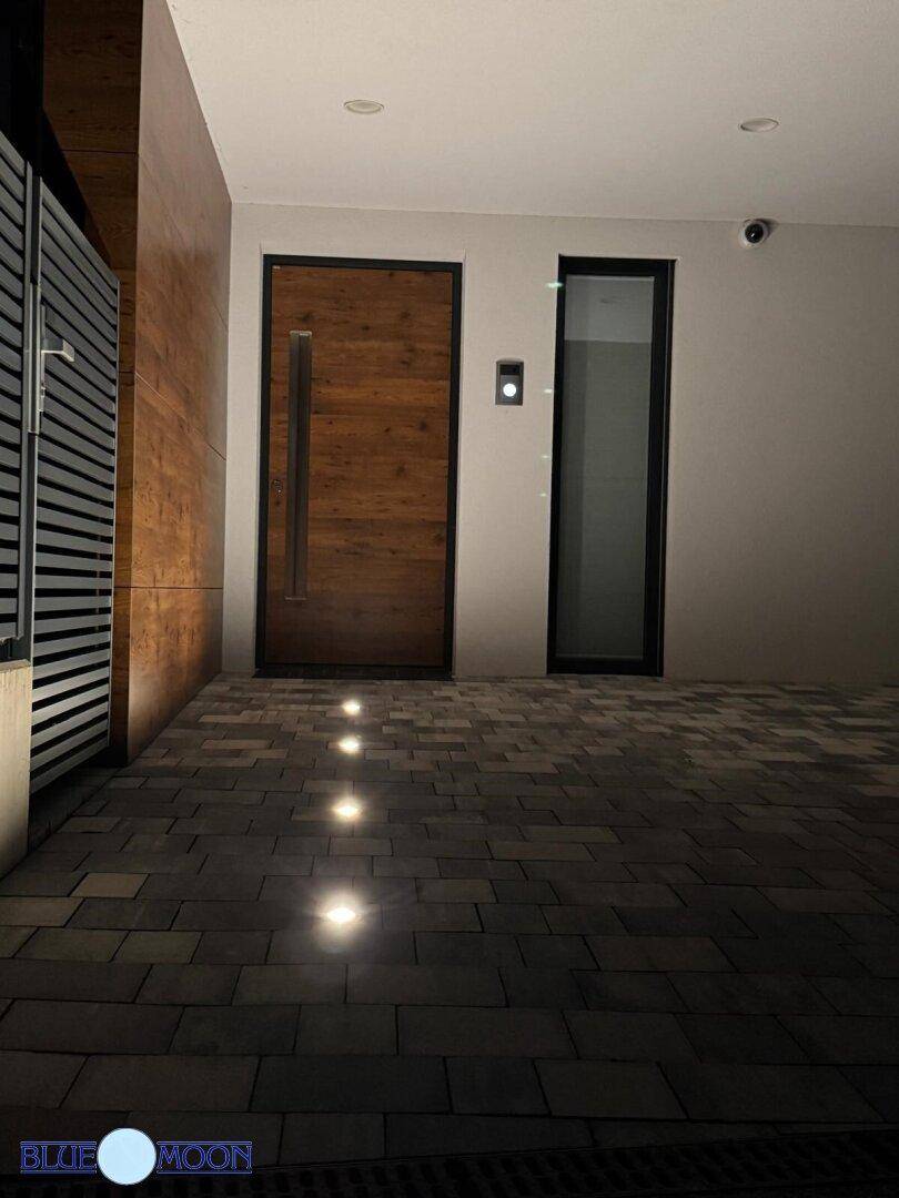 Eingangsbereich des Hauses bei Nacht mit Holztür, Kamera und gepflastertem Boden mit Lichtern.