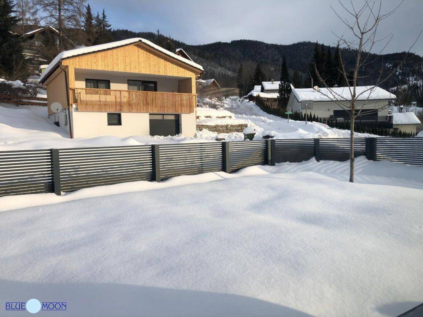 Winterlandschaft mit verschneitem Garten und einem traditionellen Holzhaus im Hintergrund.