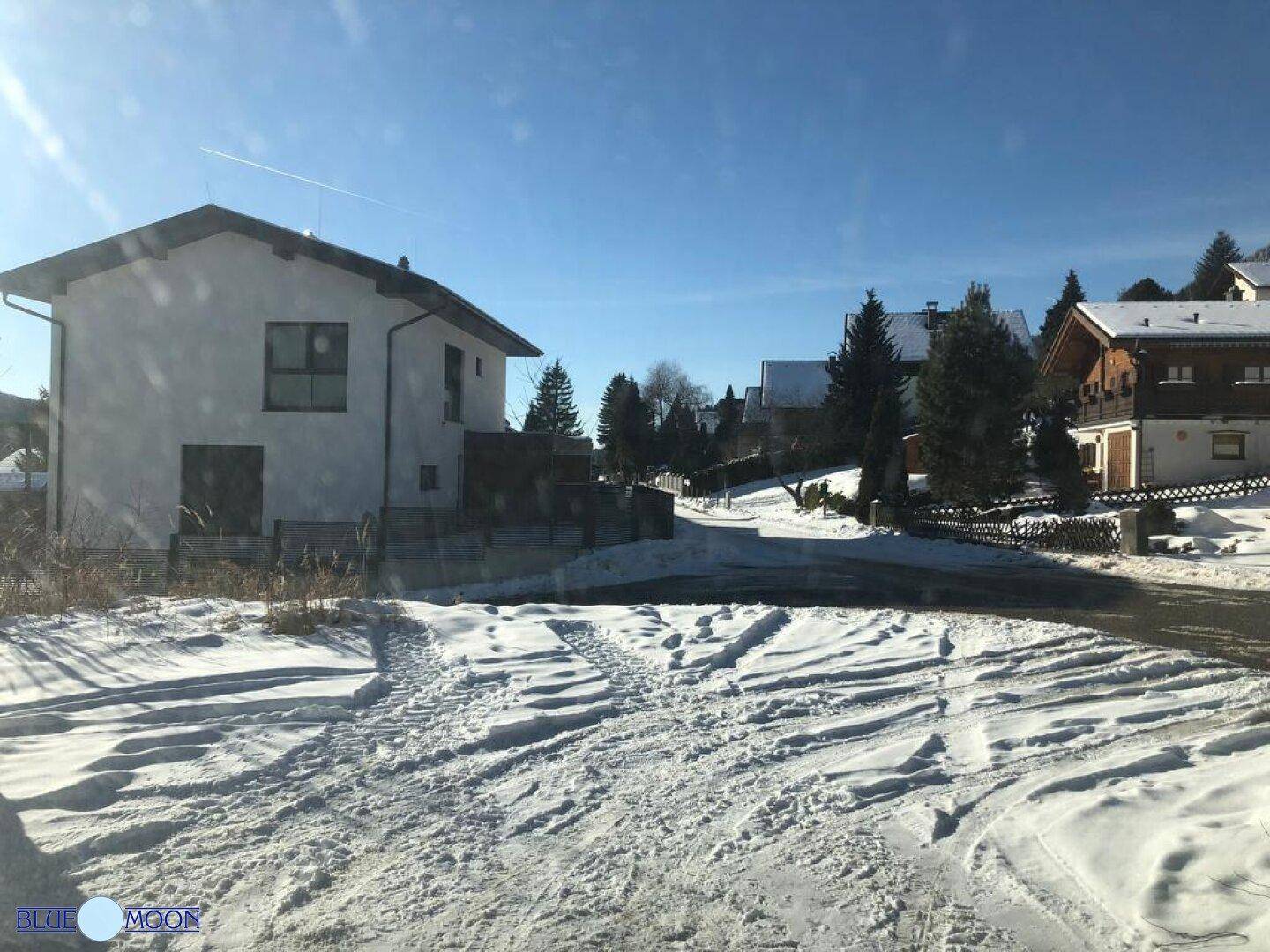 Verschneite Straße und Umgebung mit einem modernen Haus im Hintergrund an einem sonnigen Wintertag.