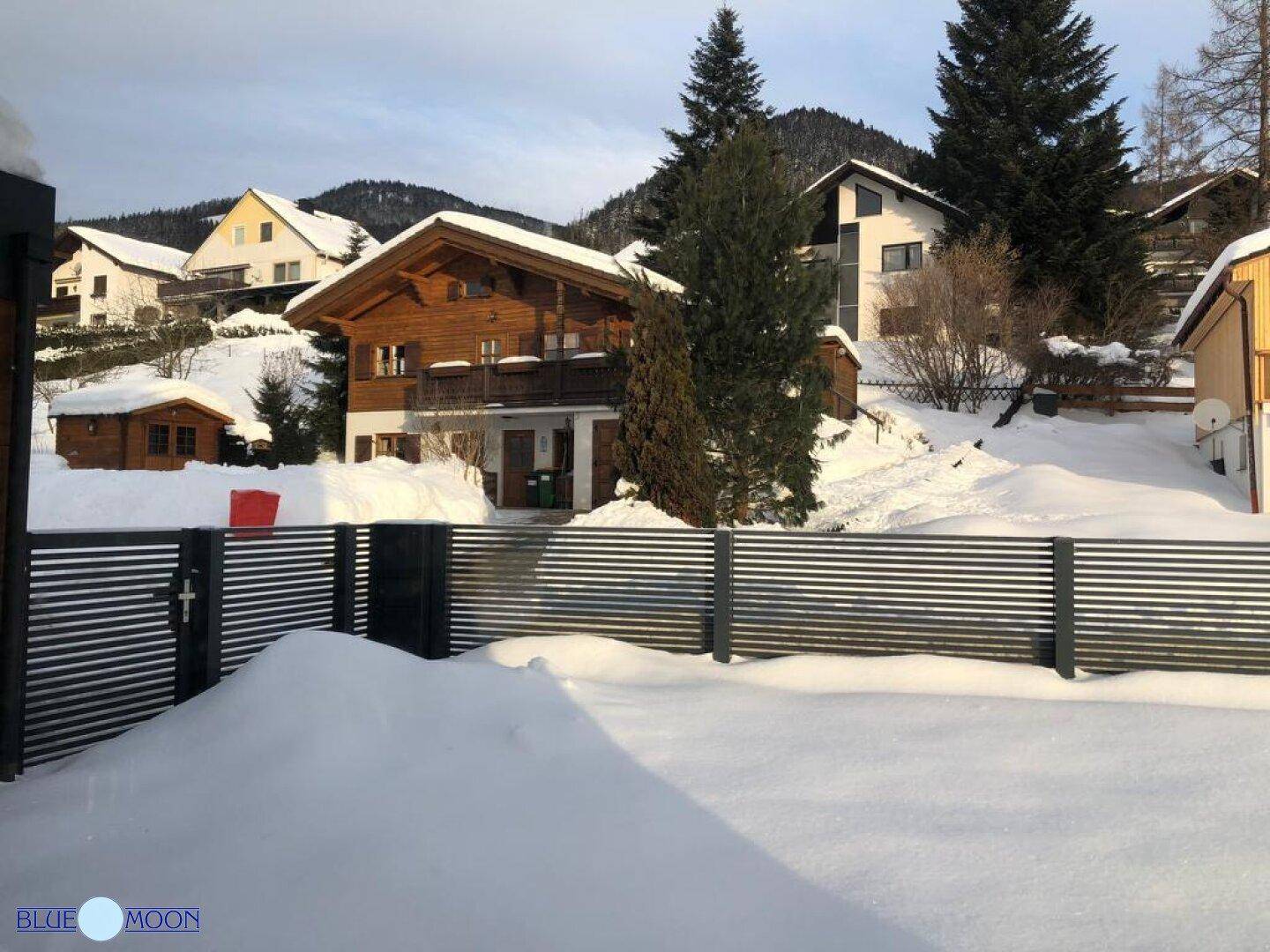 Winterlandschaft mit verschneitem Garten und einem traditionellen Holzhaus in den Bergen.