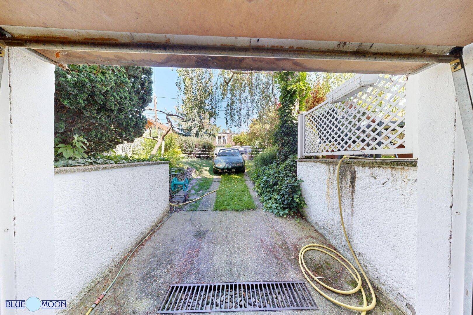 Blick aus der Garage auf die Einfahrt mit einem geparkten Auto und grüner Umgebung.