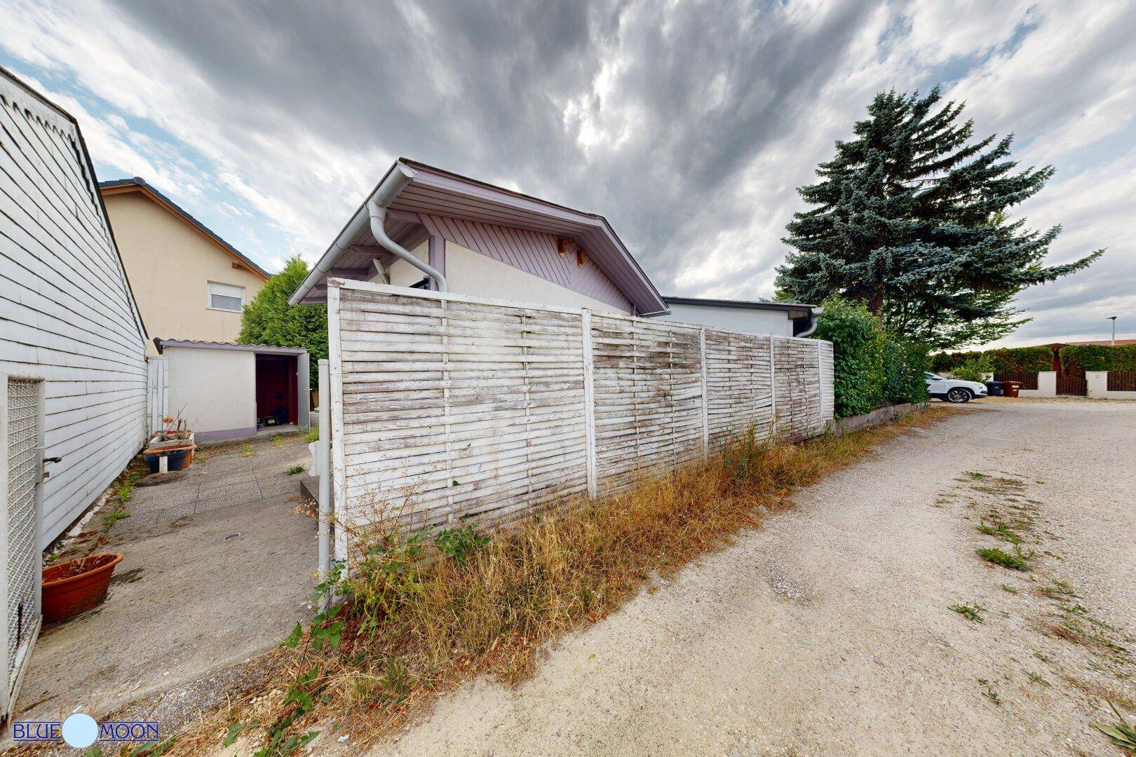 Außenansicht eines Hauses mit Holzzaun, kleinem Schuppen und Schotterweg unter bewölktem Himmel.