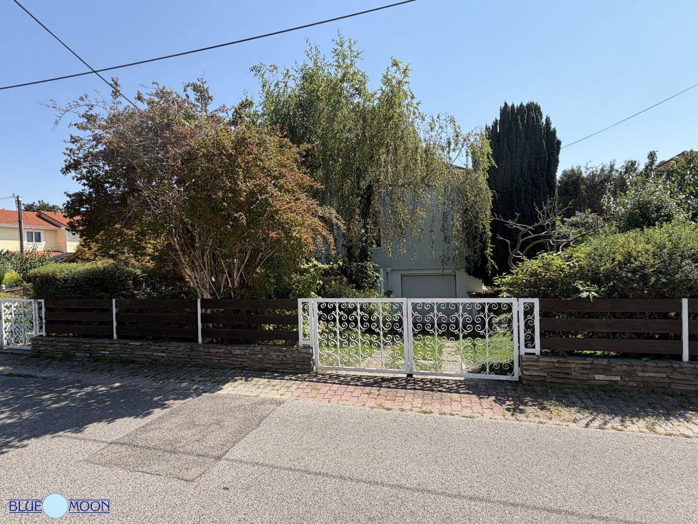 Das Haus von der Straße aus gesehen mit einem dekorativen weißen Eingangstor und einem grünen Vorgarten.