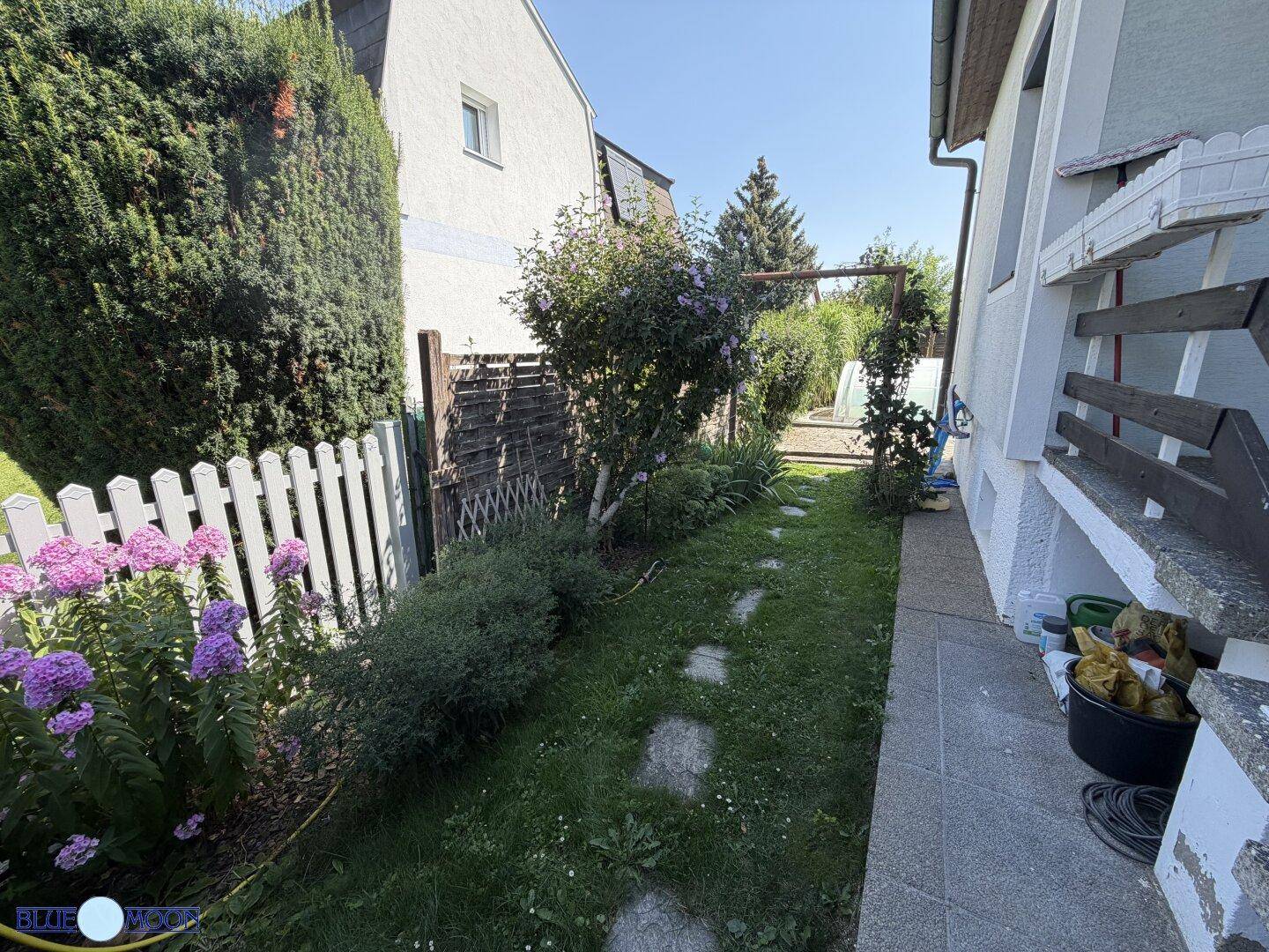Ein schmaler Gartenweg zwischen zwei Häusern, gesäumt von einem weißen Zaun und blühenden Blumen.