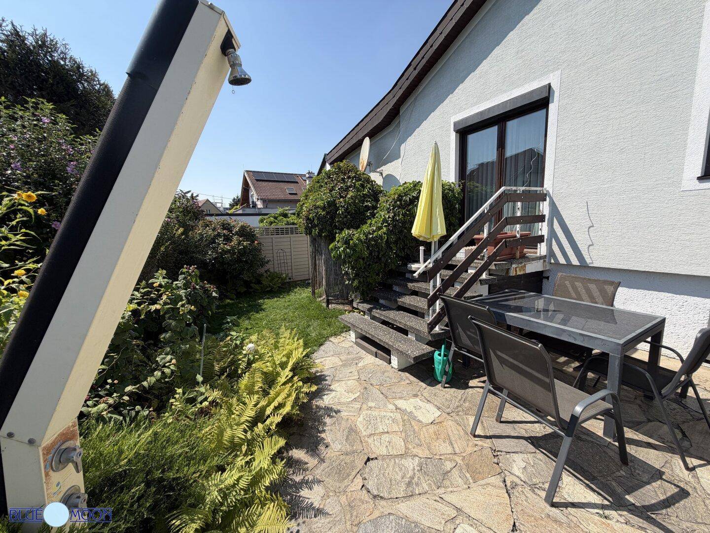 Gemütliche Steinterrasse mit Esstisch, Stühlen und einer Außendusche, ideal für Mahlzeiten im Freien.