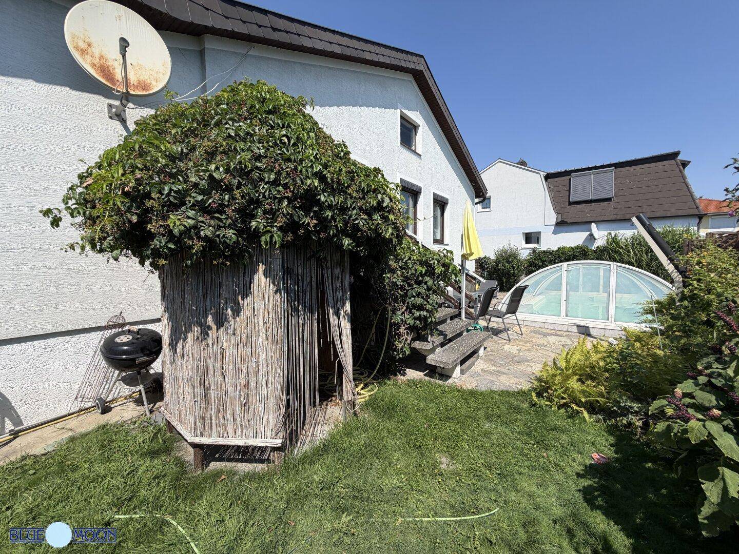 Grüner Gartenbereich mit einer von Pflanzen bewachsenen Hütte, einem Grill und Stufen zur Terrasse.