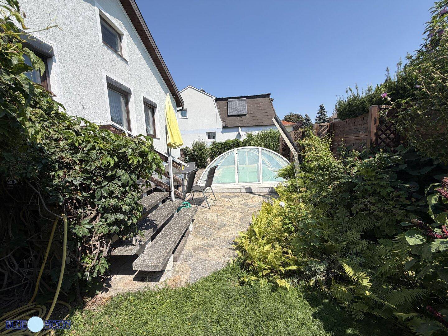 Sonniger Gartenbereich mit Stufen, die zu einer Terrasse führen, und einem überdachten Swimmingpool im Hintergrund.