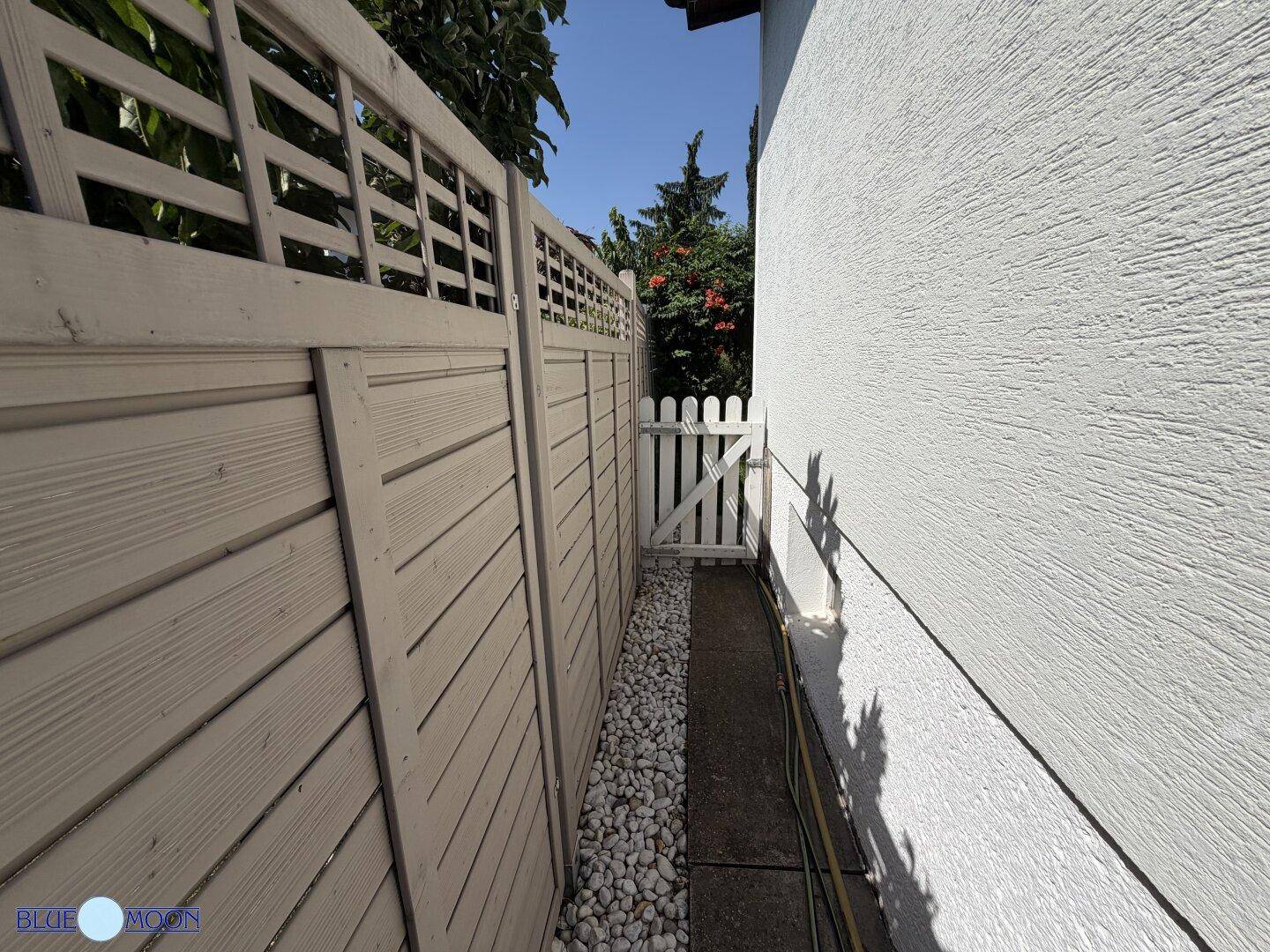 Schmaler Kiesweg zwischen der Hauswand und einem hohen Holzzaun, der zum Garten führt.