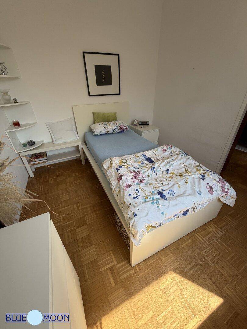 Helles Schlafzimmer mit Einzelbett, schlichten Möbeln und Parkettboden, durchflutet von Sonnenlicht.