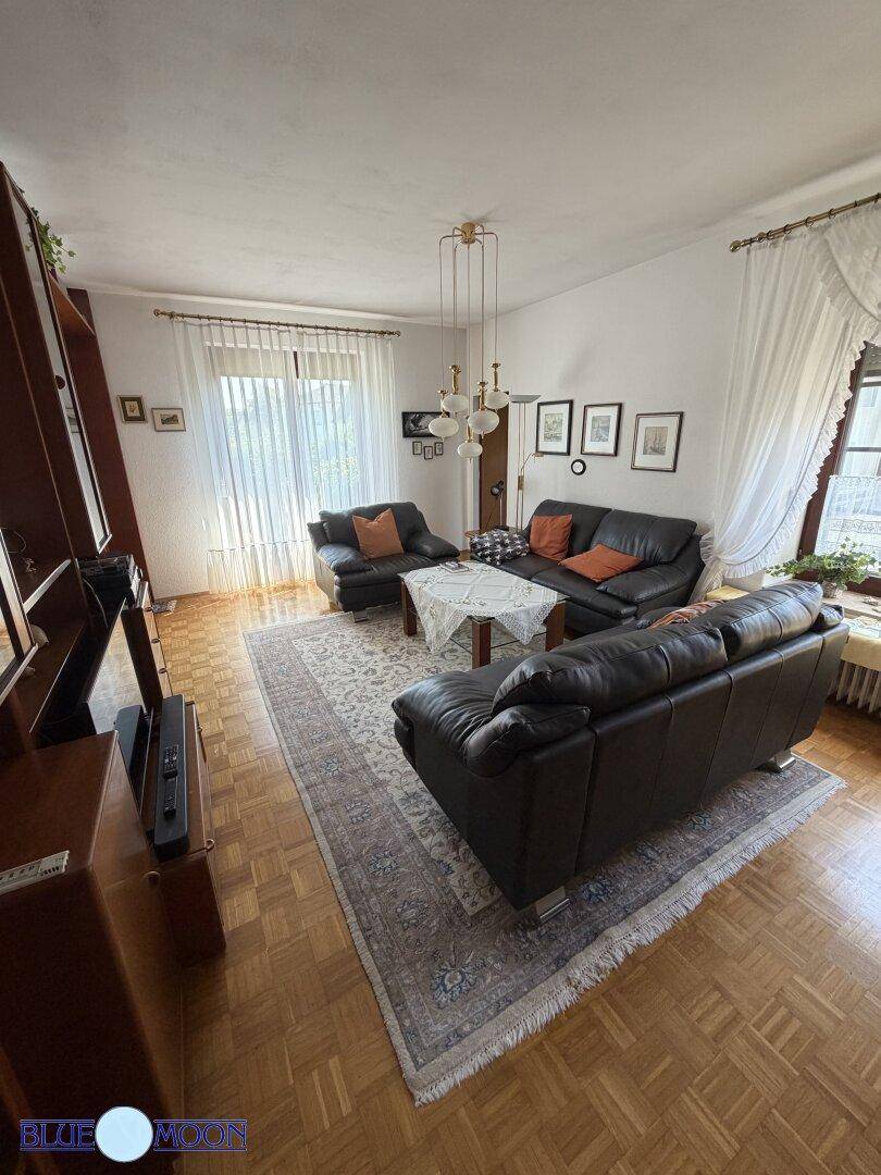 Gemütliches Wohnzimmer mit dunklen Ledersofas, Teppich und großen Fenstern mit hellen Vorhängen.