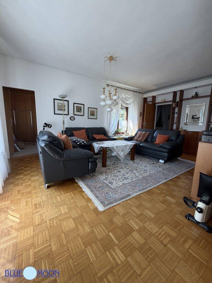 Geräumiges Wohnzimmer mit dunklen Ledersofas, Teppich und viel Tageslicht durch die Fenster.