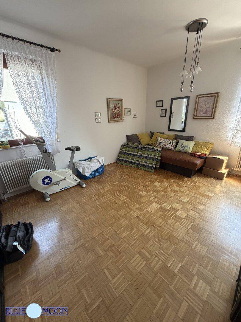 Geräumiges Zimmer mit Parkettboden, viel Tageslicht und einem Heimtrainer am Fenster.