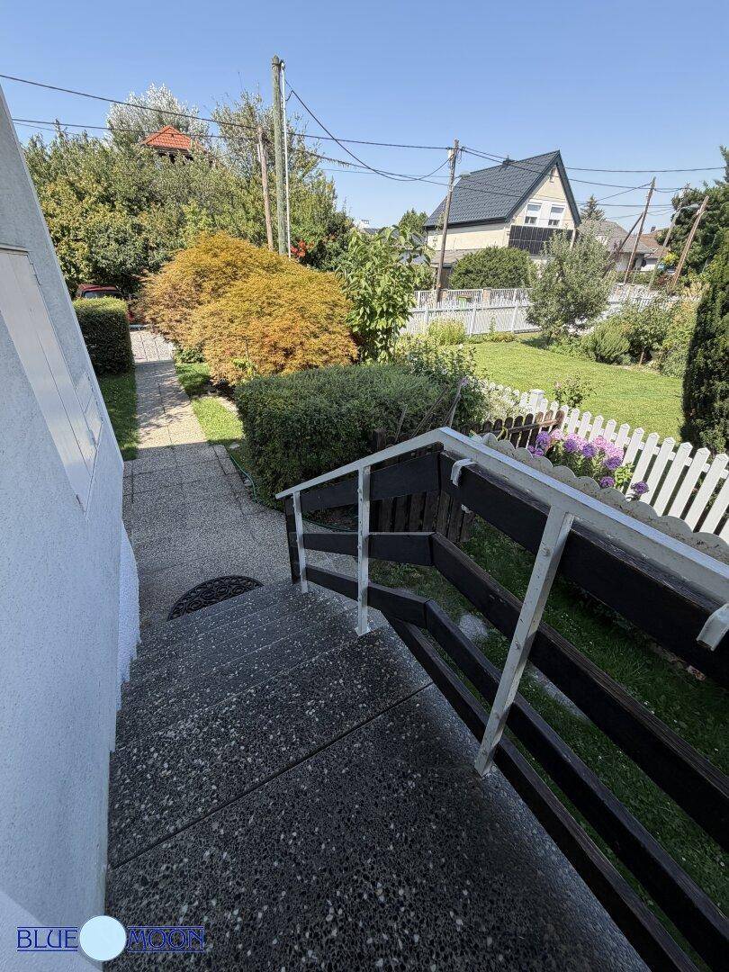Außentreppe führt hinunter in einen gepflegten Garten mit weißem Zaun und Bäumen.