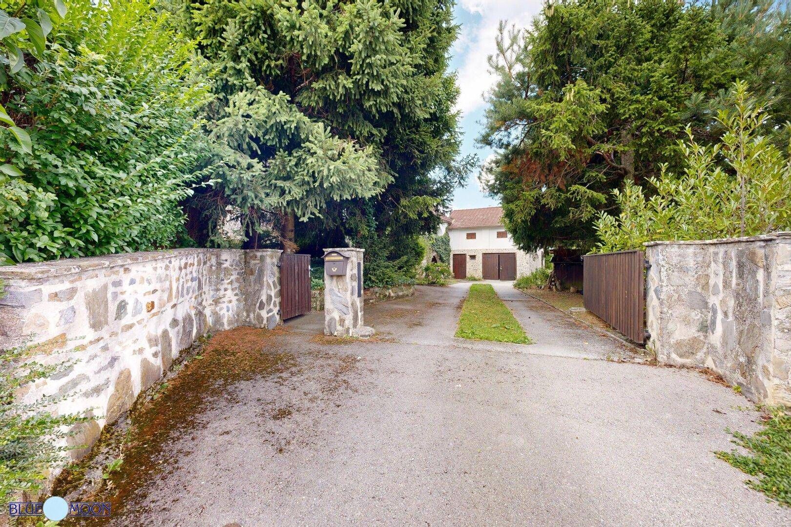 Einfahrt zum Anwesen mit Steinmauern, Holztoren und einem Briefkasten, umgeben von Bäumen.