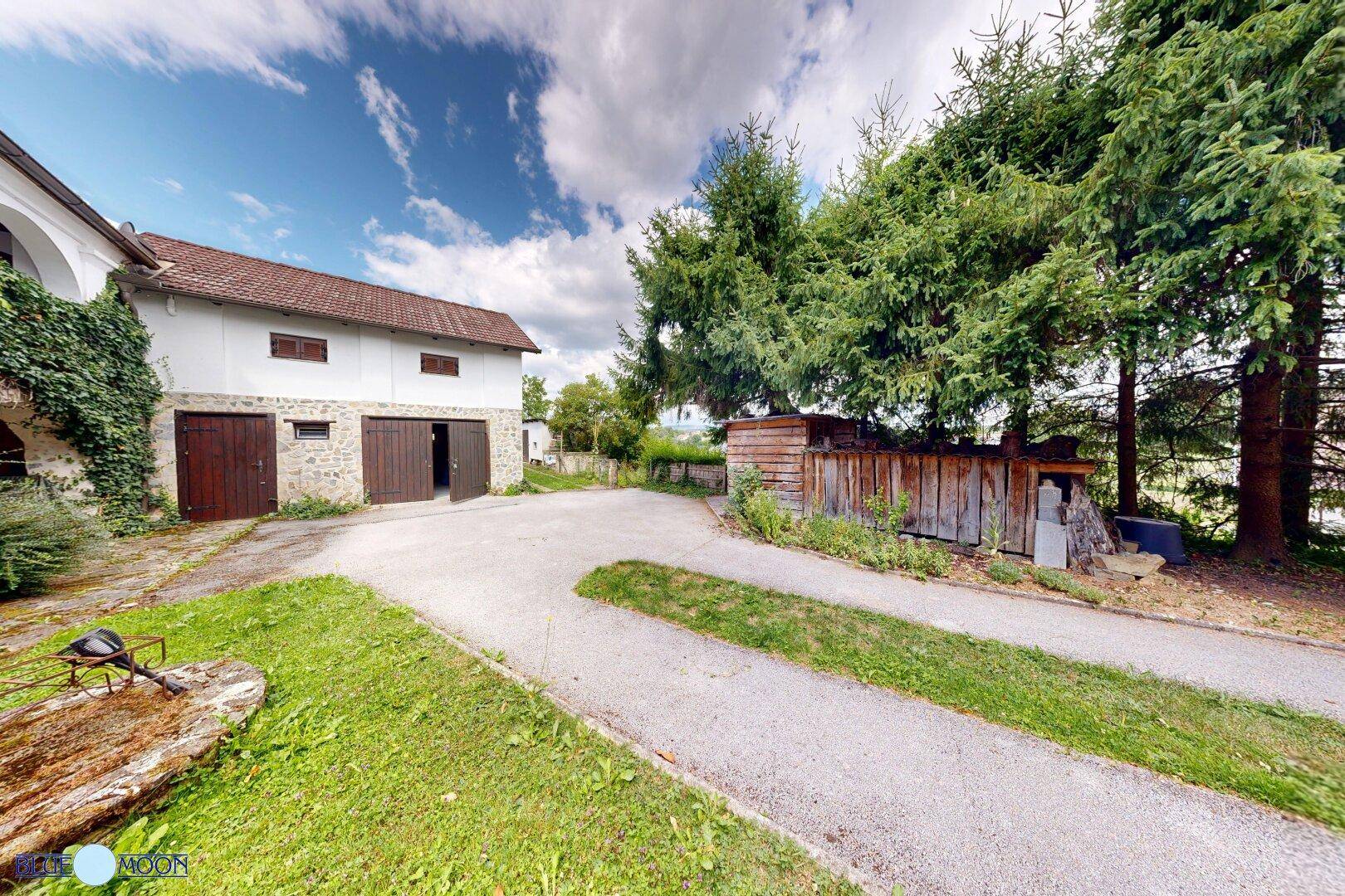 Ansicht des Nebengebäudes mit Garagentoren und einem Holzschuppen, umgeben von hohen Bäumen.