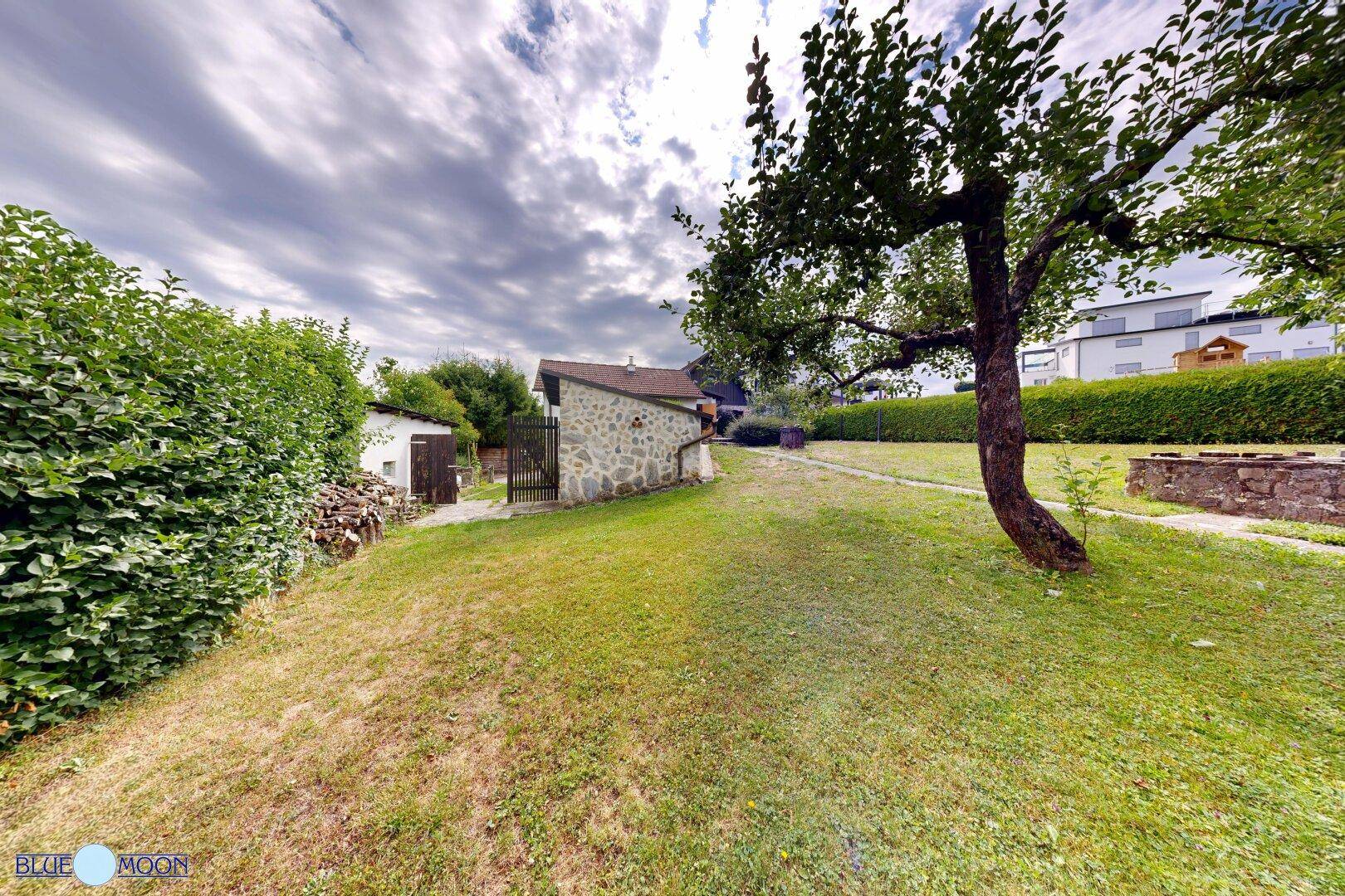 Garten mit Rasenfläche und Bäumen, im Hintergrund sind ein Steingebäude und ein Schuppen sichtbar.