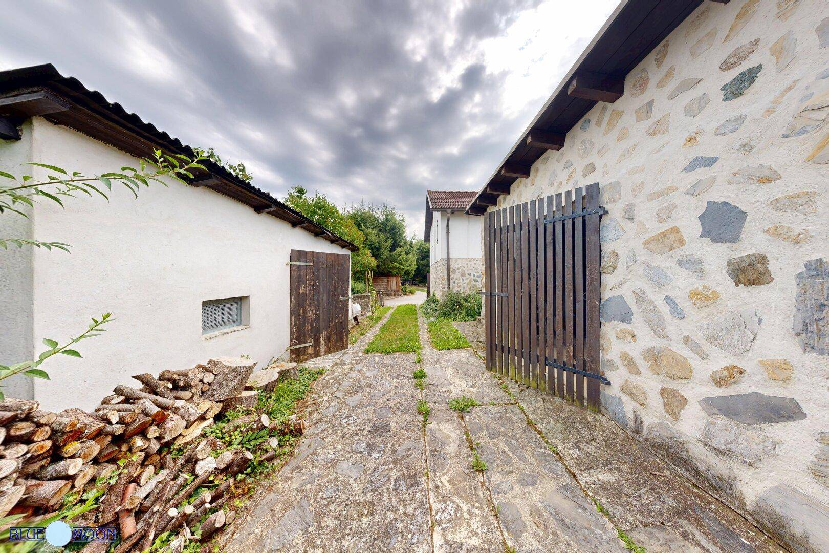 Schmaler Weg zwischen zwei rustikalen Gebäuden, eines davon mit Natursteinfassade und Holzstapel.