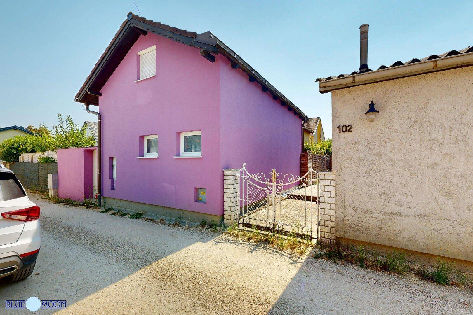 Auffälliges lila Haus mit weißem Zaun und Einfahrt, daneben ein weiteres Gebäude.