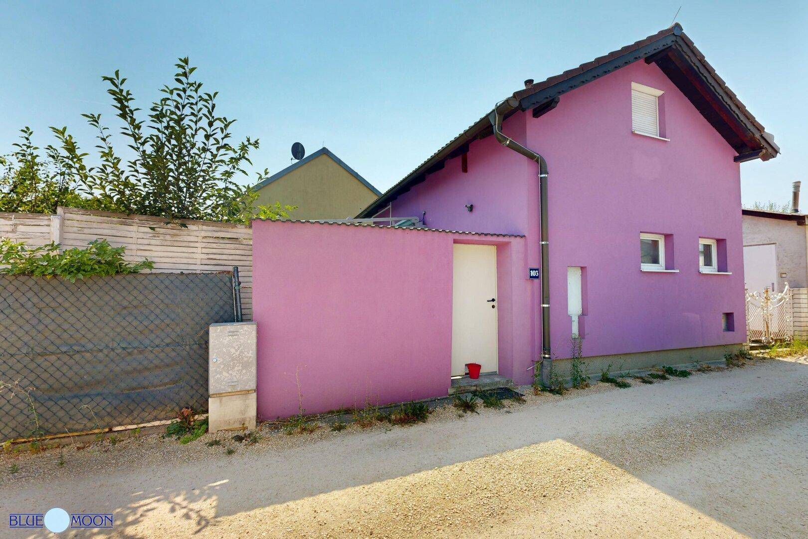 Seitenansicht des lila Hauses mit Eingangstür und Regenrinne, umgeben von einem Zaun.