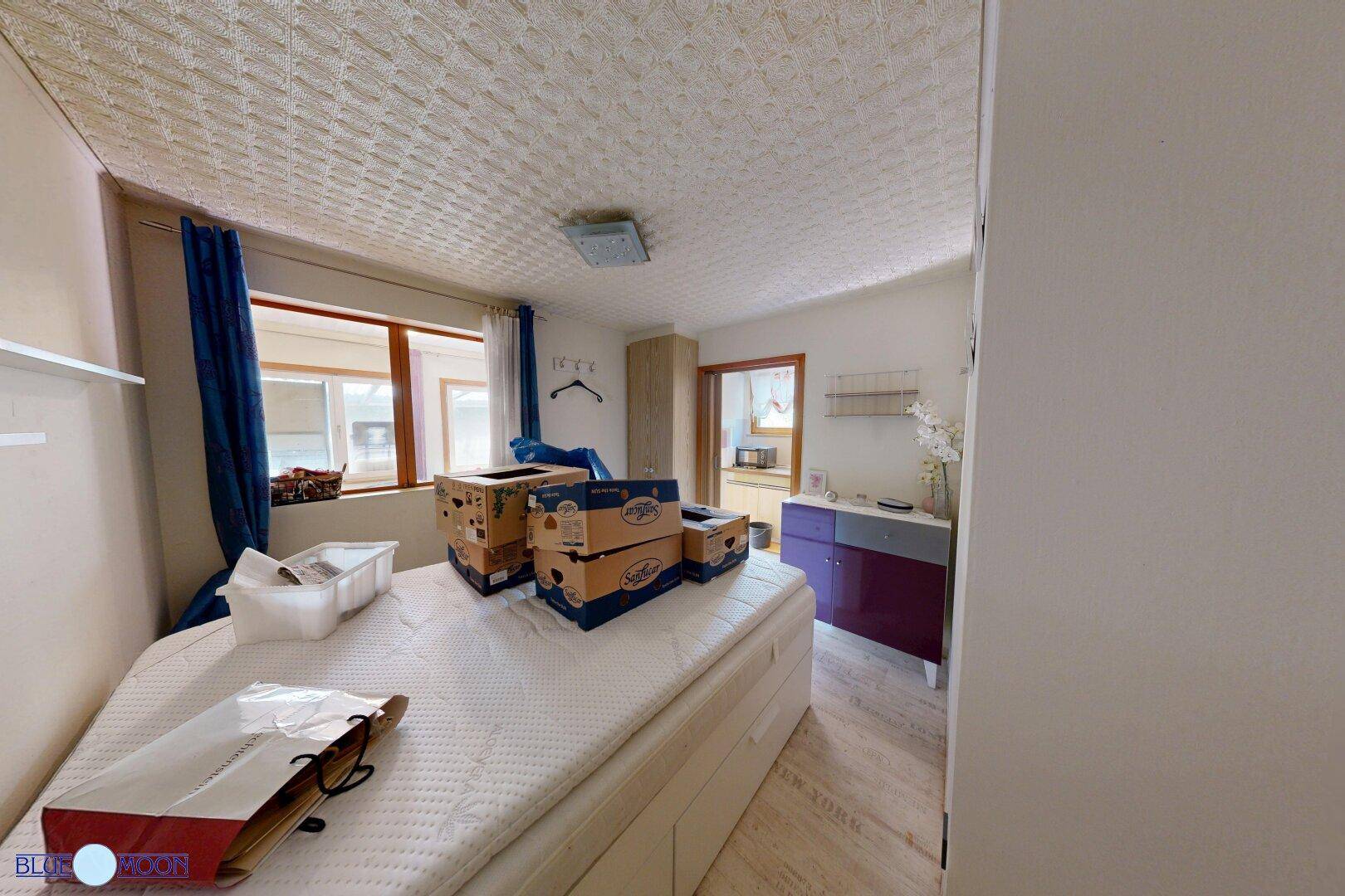Schlafzimmer mit Matratze auf dem Boden, blauen Vorhängen und Blick nach draußen.