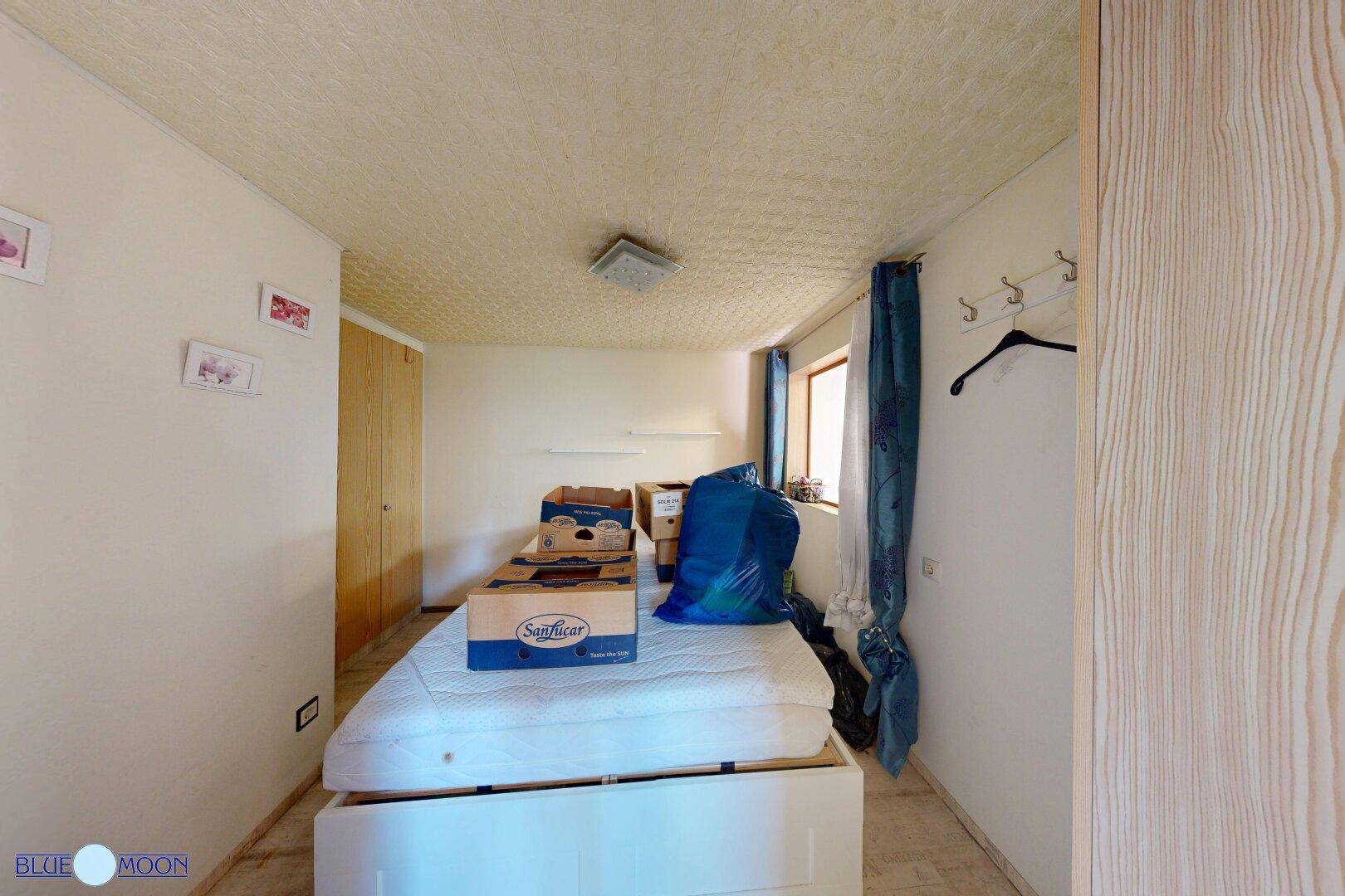 Einfaches Schlafzimmer mit Matratze, Kleiderschrank und blauen Vorhängen, hell und funktional.