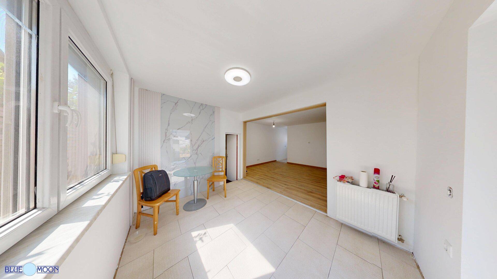 Lichtdurchfluteter Raum mit Fliesenboden, Fensterfront und einem kleinen Tisch.