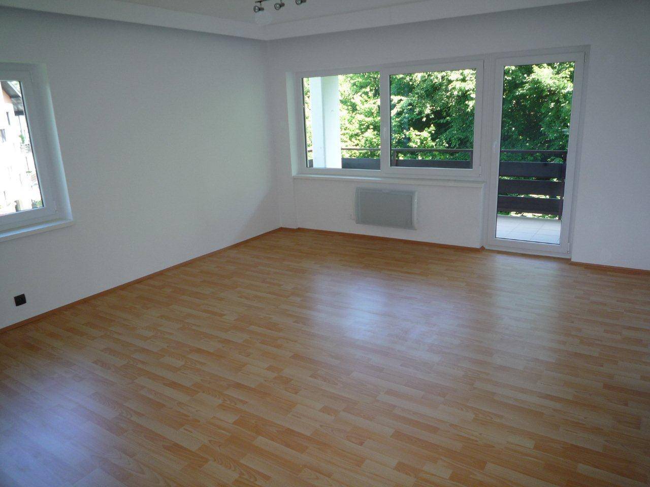 Helles, leeres Wohnzimmer mit Laminatboden, weißen Wänden und Zugang zu einem Balkon.