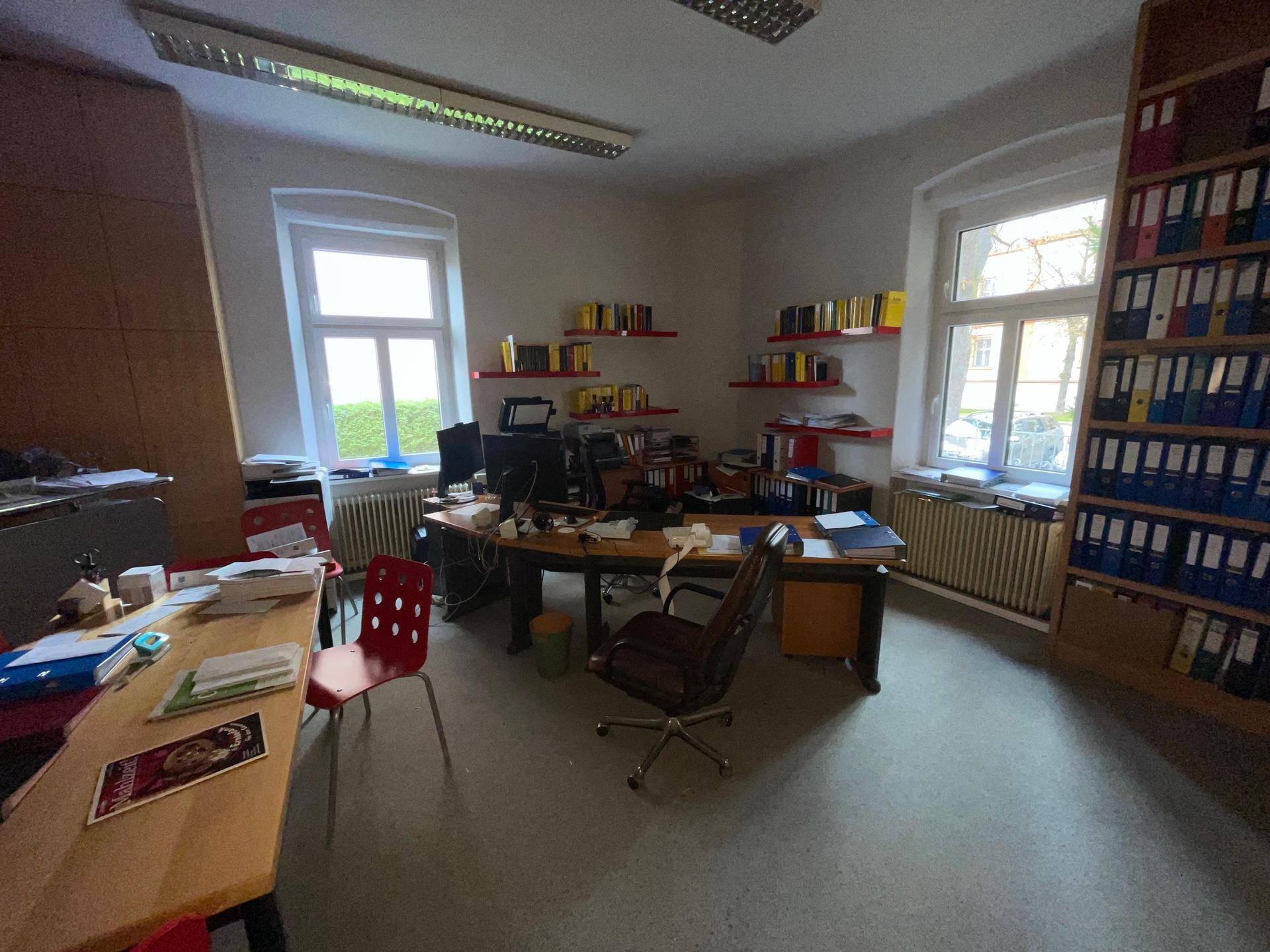 Helles Büro mit zwei Fenstern, Holzschreibtisch und Regalen voller Aktenordner, gut beleuchtet.