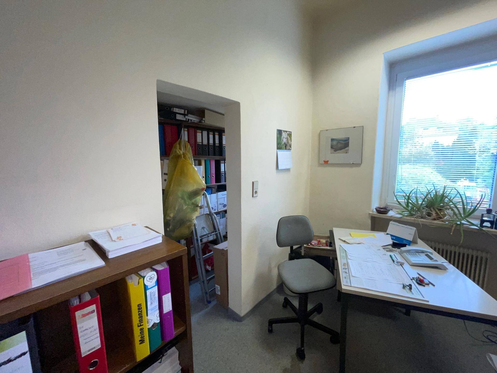 Kleiner Bürobereich mit Schreibtisch am Fenster und einem Durchgang zu weiteren Aktenregalen.