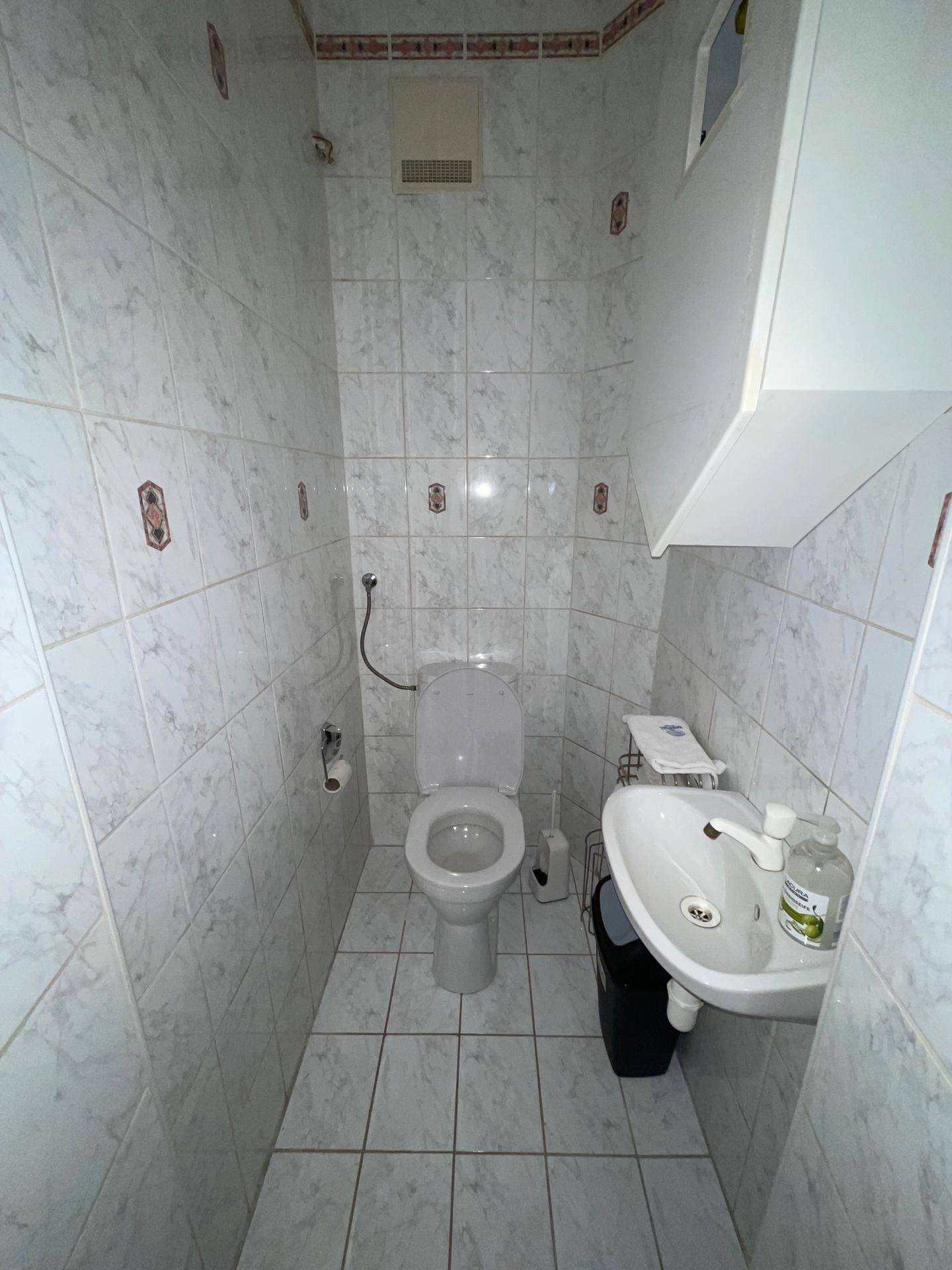 Schmale Toilette mit weißen Fliesen, WC und kleinem Waschbecken, einfach und sauber.