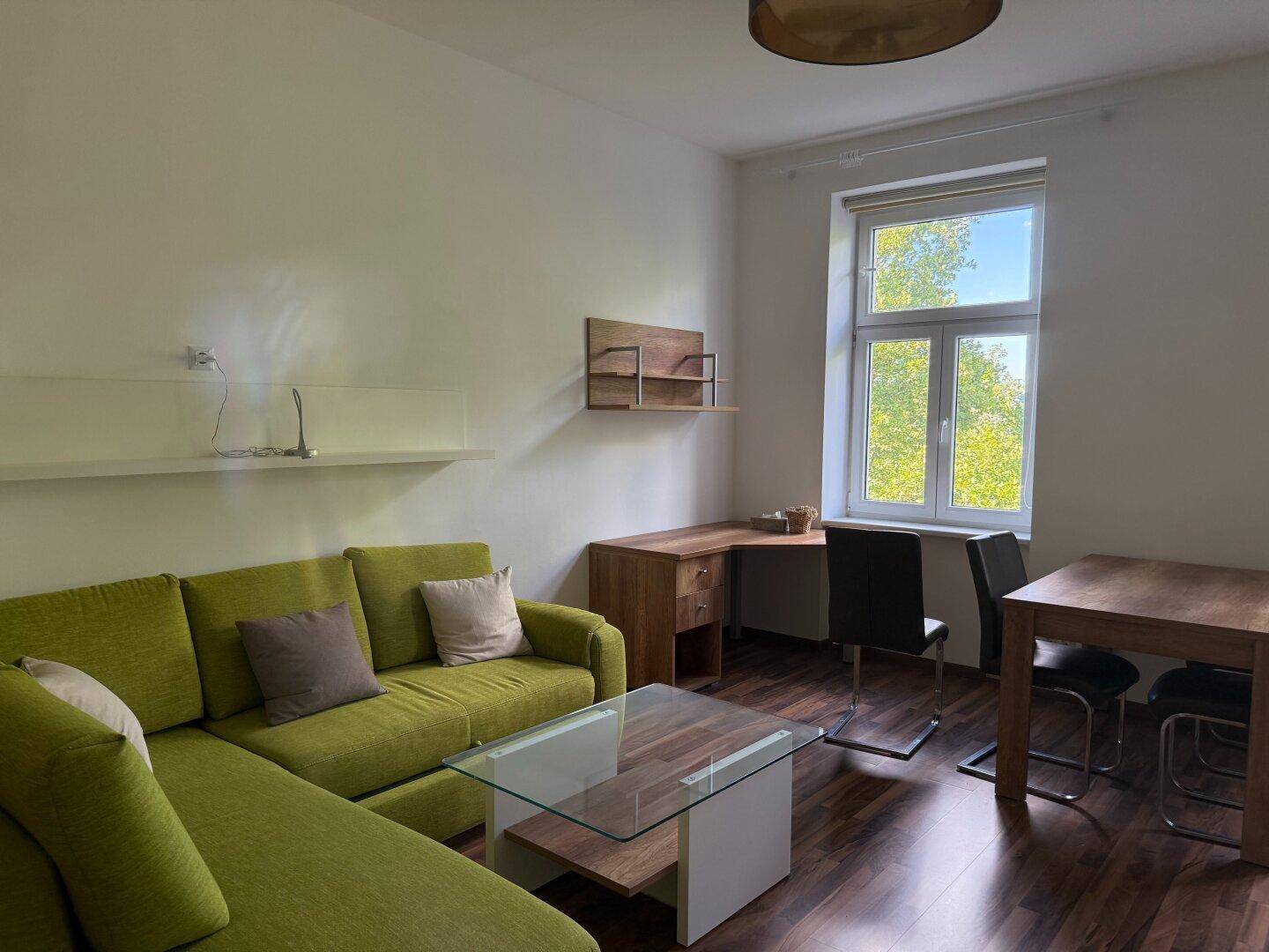 Gemütlicher Wohnbereich mit L-förmigem Sofa, Schreibtisch und viel Tageslicht.