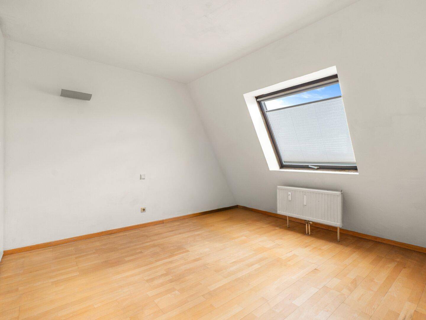 Ruhiges Zimmer mit Holzfußboden, weiß gestrichenen Wänden und einem Dachfenster mit Rollo.