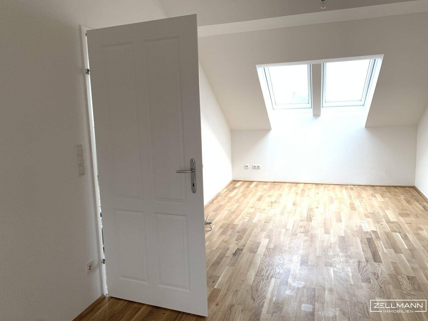 Helles Dachgeschosszimmer mit Holzboden und zwei großen Dachfenstern, durch eine weiße Tür zugänglich.
