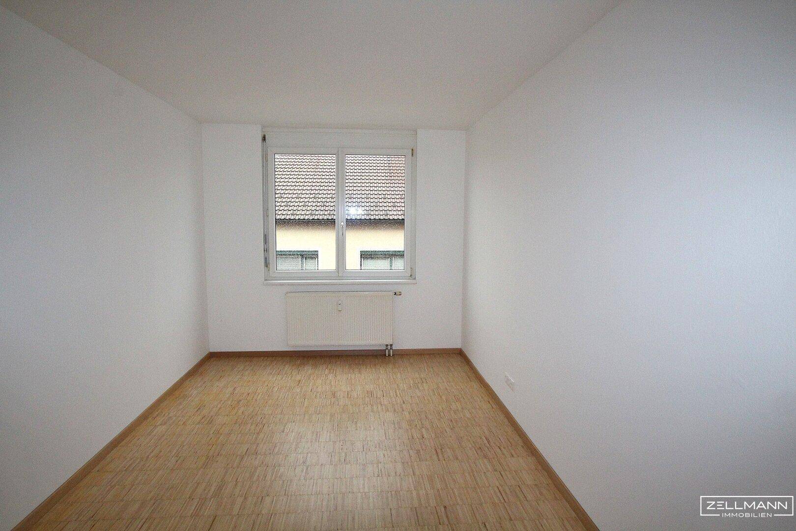 Ruhiges Schlafzimmer mit Parkettboden, weißen Wänden und einem Fenster mit Rollladen für Privatsphäre.