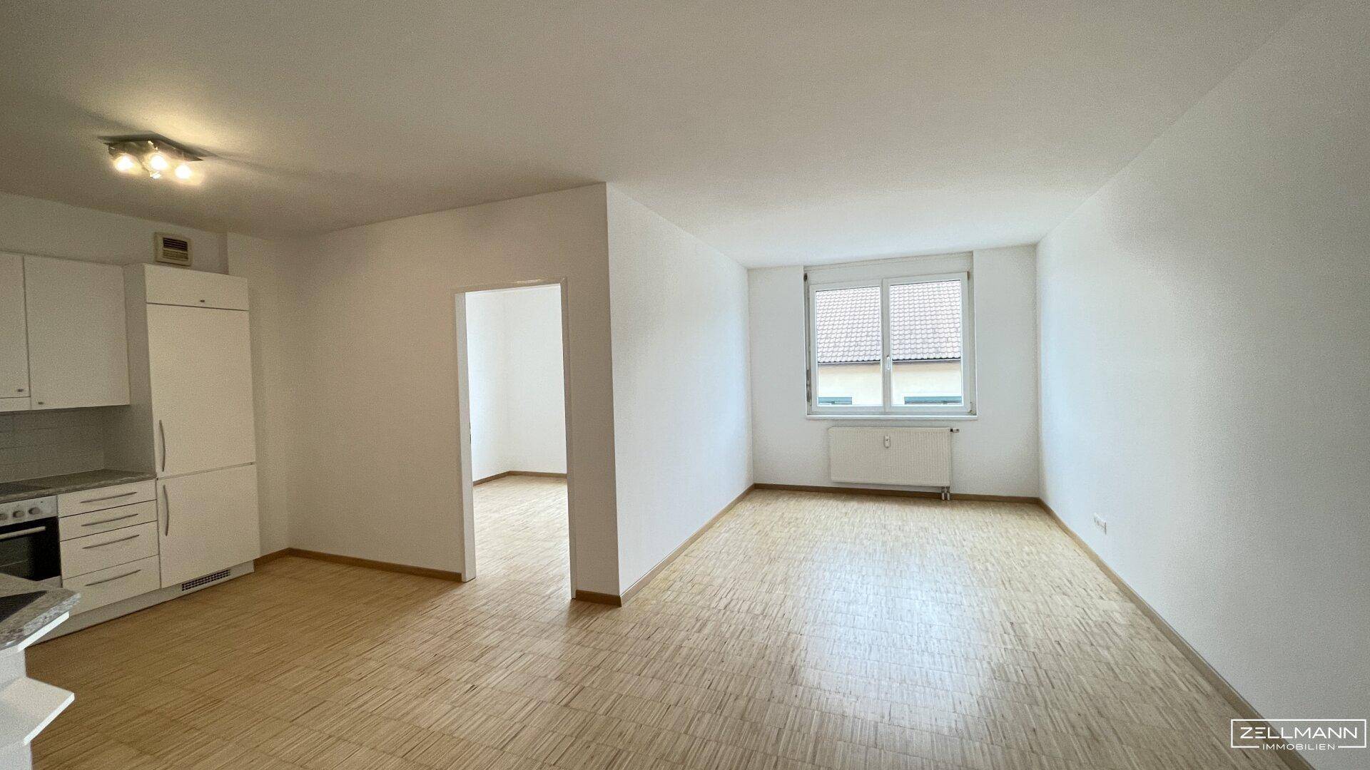 Offener Wohnbereich mit integrierter Küche, Parkettboden und einem Fenster mit Tageslicht.
