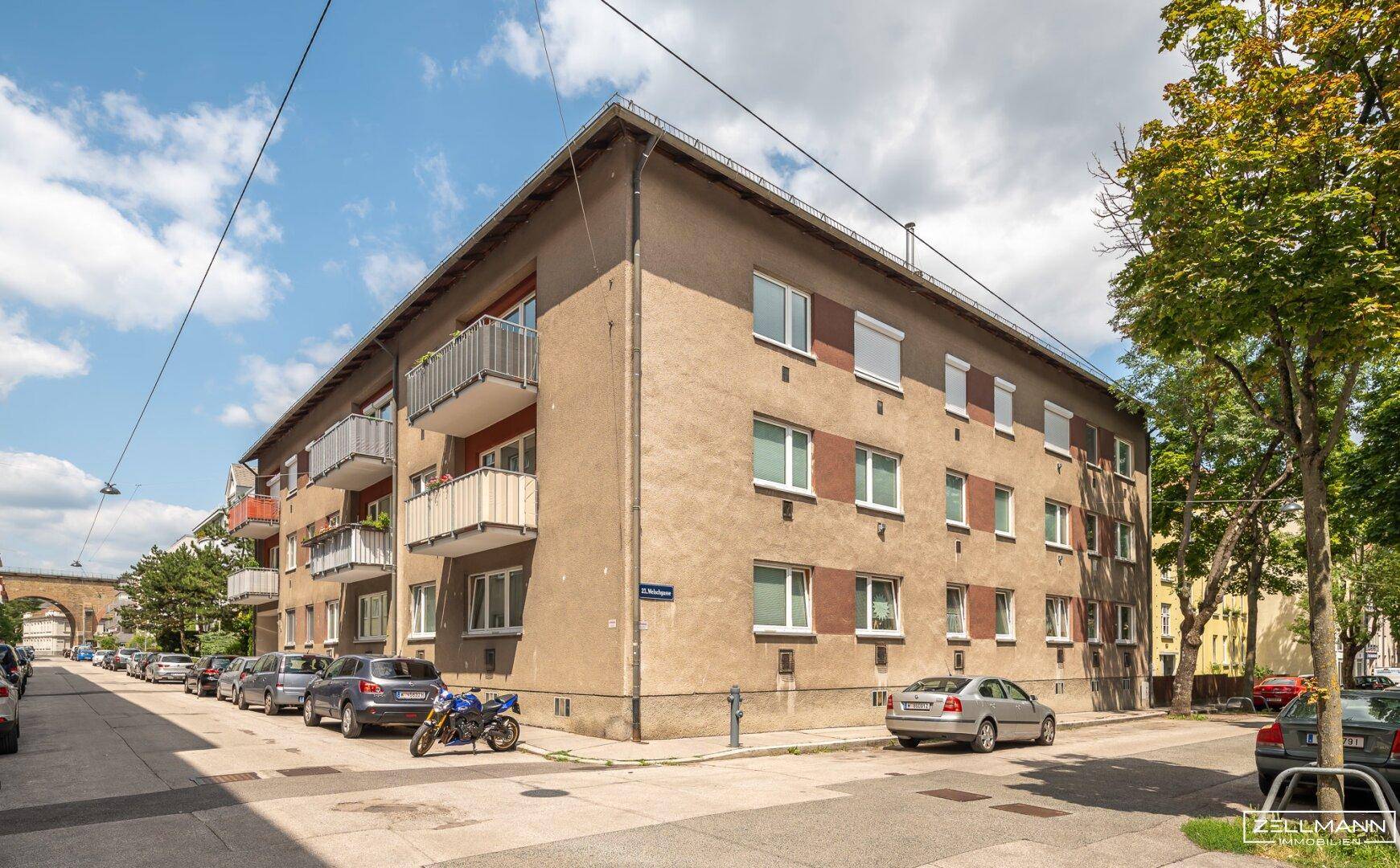 Mehrfamilienhaus mit Balkonen an einer sonnigen Straße, umgeben von Bäumen.